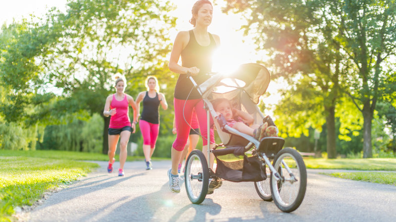 A woman jogs on a sunny day in a park, pushing a stroller with a child inside. Other joggers are visible in the background. The park is lush with green trees and grass, and sunlight filters through the leaves, creating a bright and vibrant atmosphere.