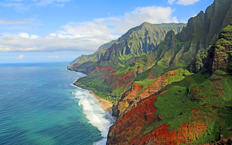 A scenic view of the Nā Pali Coast in Hawaii, featuring rugged green cliffs with red and brown hues, cascading towards the blue ocean. White waves crash against the shore, and the sky is partly cloudy with patches of blue.