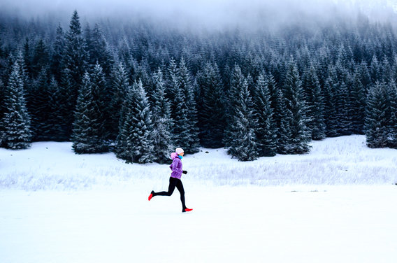 A person dressed in a purple jacket and black pants runs across a snow-covered landscape with a dense forest of snow-dusted evergreen trees in the background. Thick fog hovers above the treetops, creating a cold and serene atmosphere.