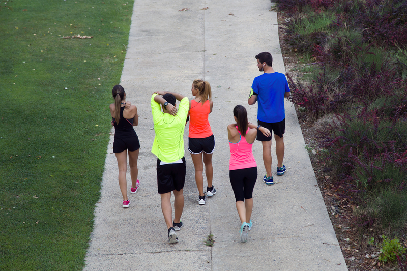 Five people wearing athletic clothing are walking on a paved path outdoors. Three women and two men are seen from behind, with some stretching their arms. On the left is a grassy area, and on the right, there are some shrubs and plants.