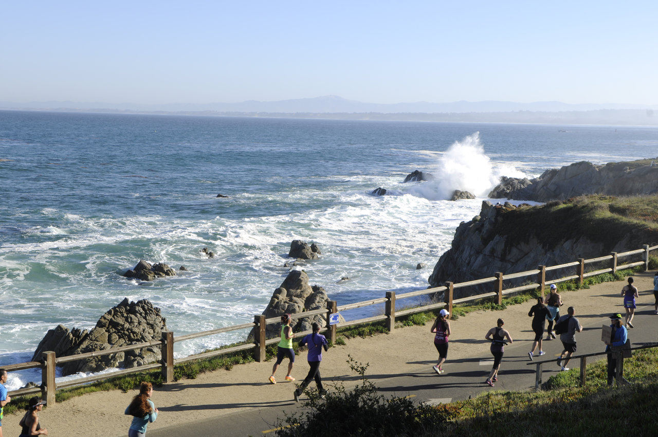 A group of people jogs on a paved path along a scenic coastline with crashing waves and rocky outcrops. The sky is clear, and the ocean stretches into the horizon. A wooden fence runs along the path, separating it from the cliffs and ocean below.