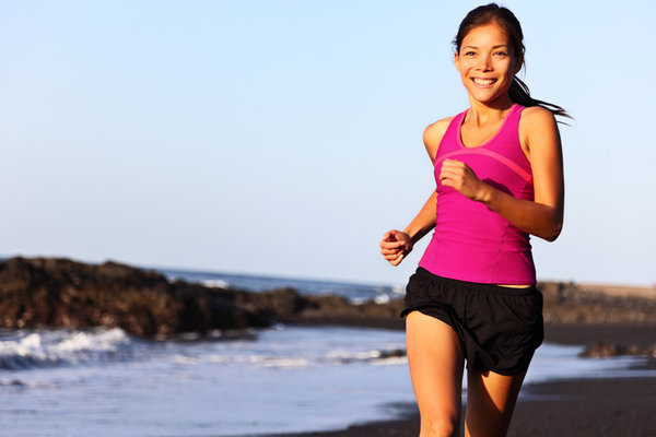 A woman in a pink tank top and black shorts is jogging along a beach. The ocean and rocky shoreline are visible in the background, and the sky is clear and blue. She is smiling and appears to be enjoying her run.