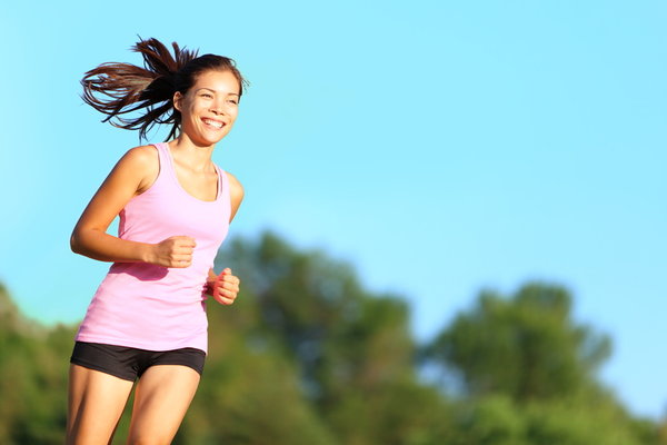 A woman wearing a pink tank top and black shorts is jogging outdoors. She has a ponytail and is smiling, with a backdrop of green trees and a clear blue sky.