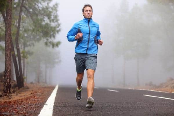 Person running on an empty road surrounded by fog and trees. The person is wearing a blue jacket, gray shorts, and black running shoes, with a determined expression. The scene is misty, creating a serene, atmospheric background.