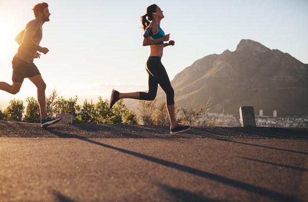 Two people, a man and a woman, are running outdoors on a sunny day. They are dressed in athletic wear with a mountainous landscape in the background. The sun is low, creating long shadows on the pavement. Greenery and city buildings are visible in the distance.