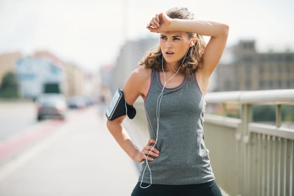 A woman dressed in athletic wear is taking a break during a run on a bridge. She is holding one hand to her forehead and an arm strap with a smartphone is attached to her other arm. Earbuds connected to the phone hang from her ears, and her expression appears tired.