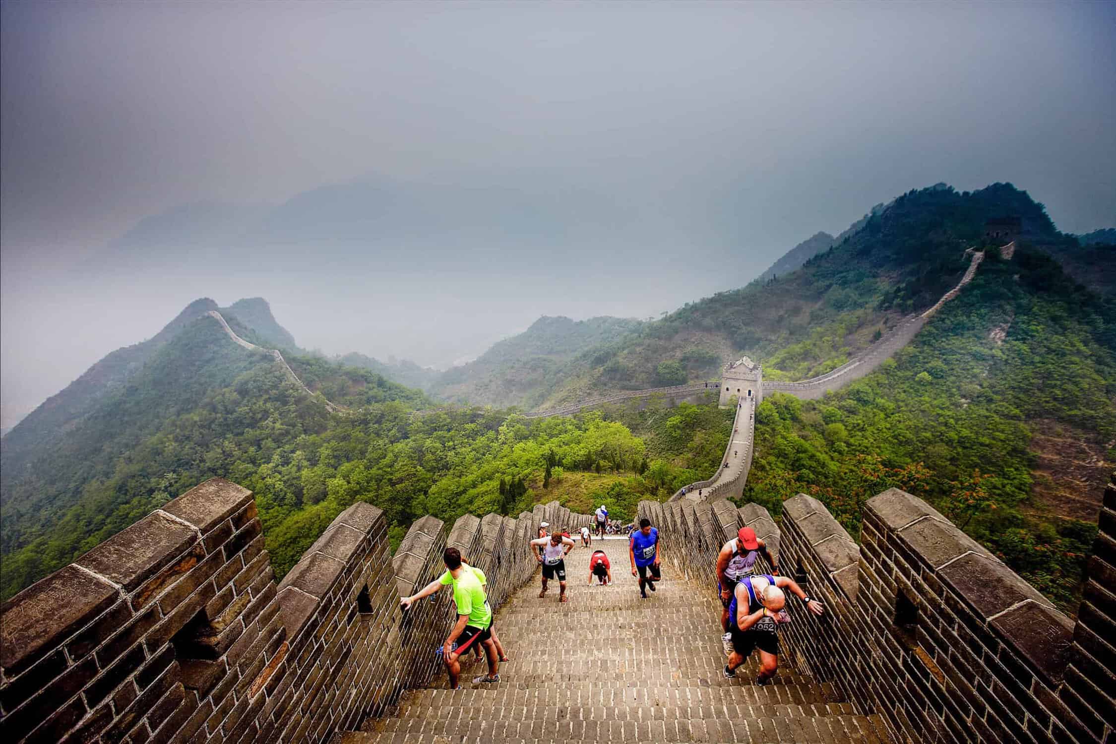 Tourists climb a steep section of the Great Wall of China, surrounded by mist and lush green hills. The ancient structure stretches into the distance, winding through the rugged landscape under a cloudy sky, reminiscent of adventurous half marathons that challenge and inspire.