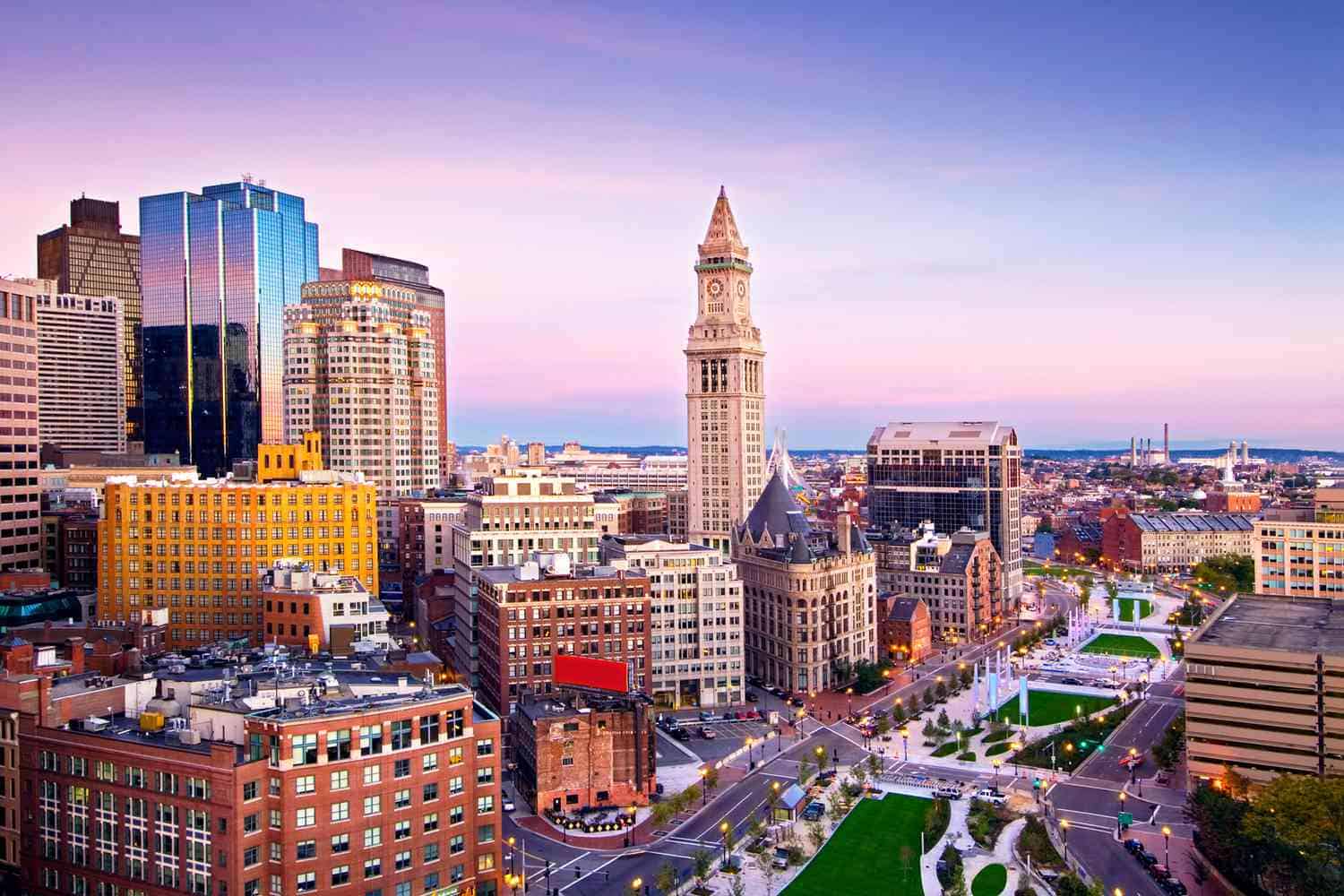 A vibrant cityscape of Boston at sunset. Prominent buildings include the Custom House Tower in the center, surrounded by a mix of modern skyscrapers and historic architecture. As runners prepare for the Boston half marathon, green spaces and busy streets are visible, with a colorful sky in the background.