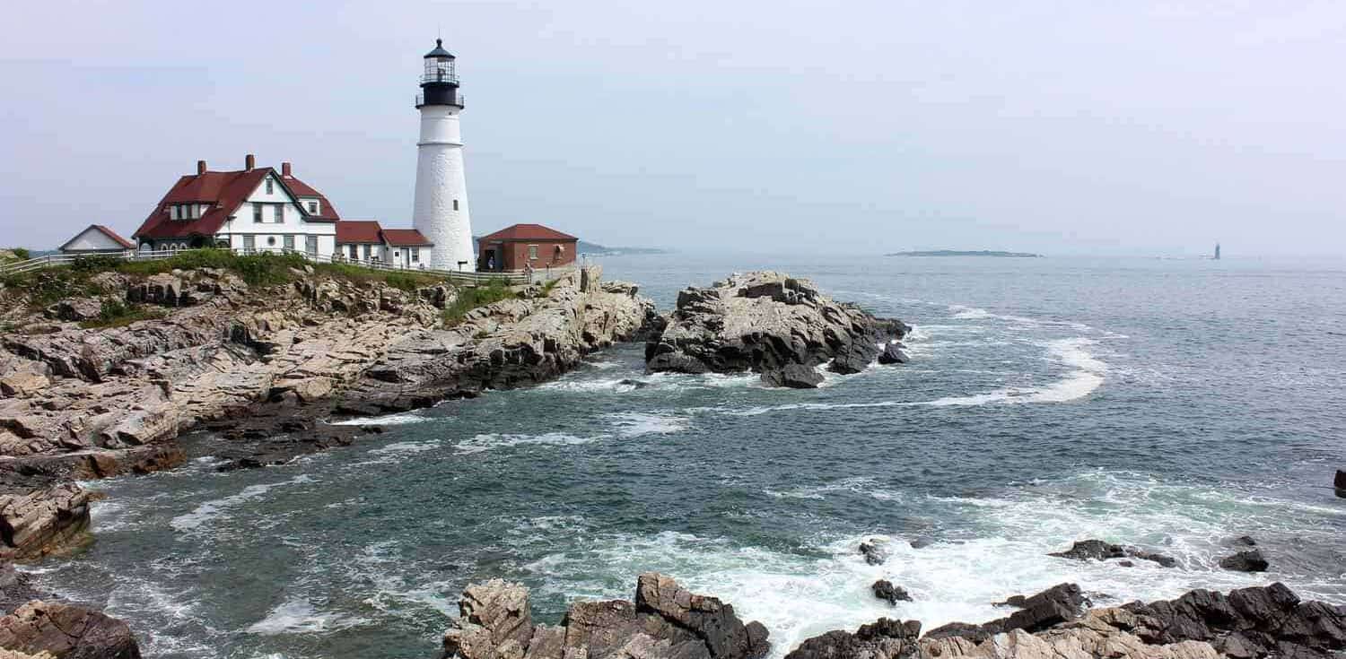 A picturesque coastal scene featuring a white lighthouse with a black top and red-roofed buildings nestled on a rocky shoreline. The ocean waves crash against the rocks under a slightly hazy sky, reminiscent of New England's scenic backdrop for half marathons. Additional rocks and a small island are visible in the distance.