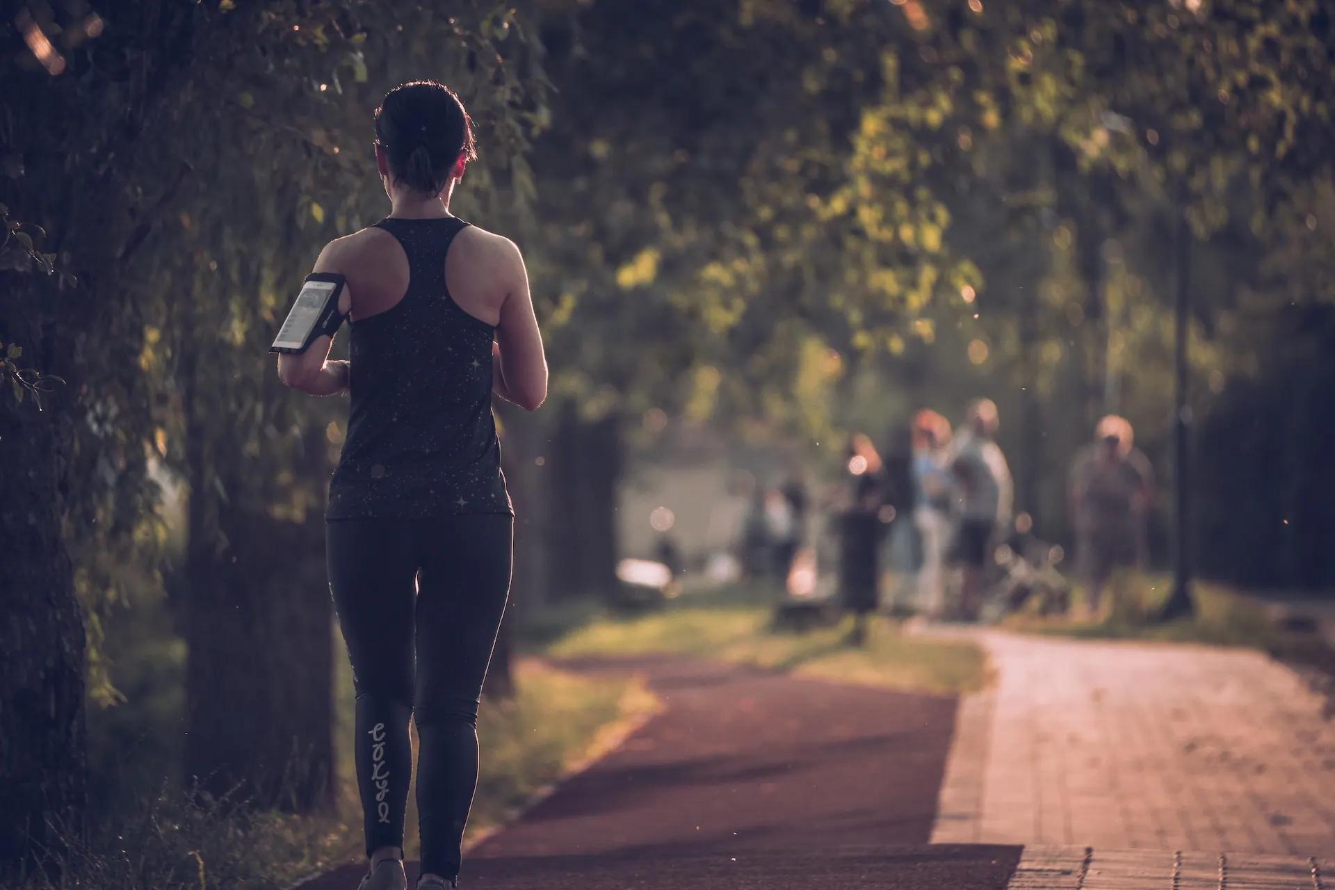 A person wearing workout clothes jogs on a shaded path, holding a smartphone in their left hand. The background is a tree-lined path with blurred figures of people and cyclists further along the trail. Sunlight filters through the leaves, casting dappled shadows.