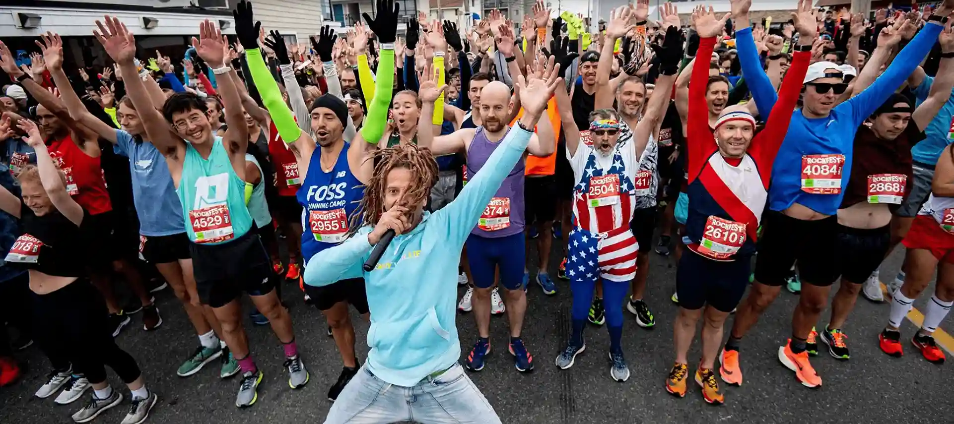 An energetic crowd of marathon runners raises their hands at the starting line. The participants wear colorful athletic gear and race bibs. A person in front with dreadlocks and a blue sweatshirt appears to be rallying the group with a microphone.